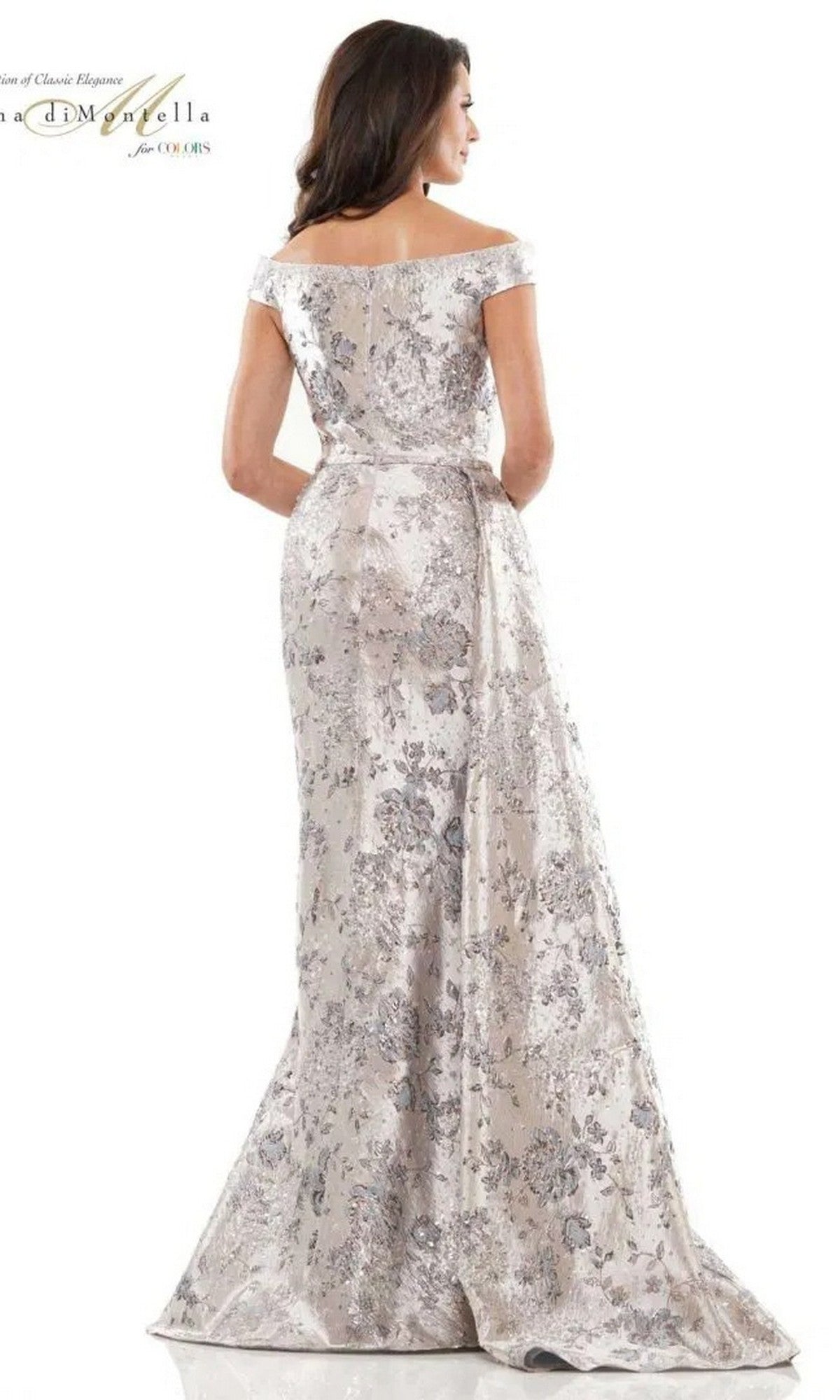 A woman faces away from the camera, wearing the Rina Di Montella RD2912 long formal dress—an off-the-shoulder, floor-length silver jacquard gown—against a plain backdrop.